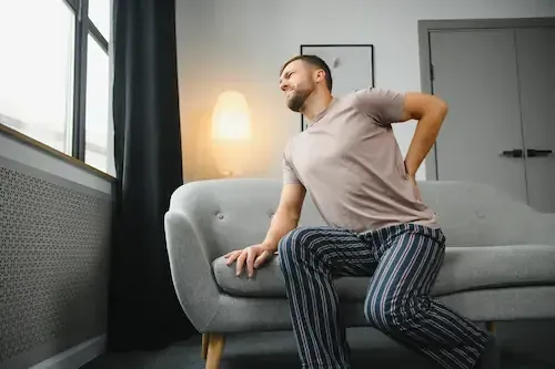 A man sitting on a sofa at home, reaching for his lower back.