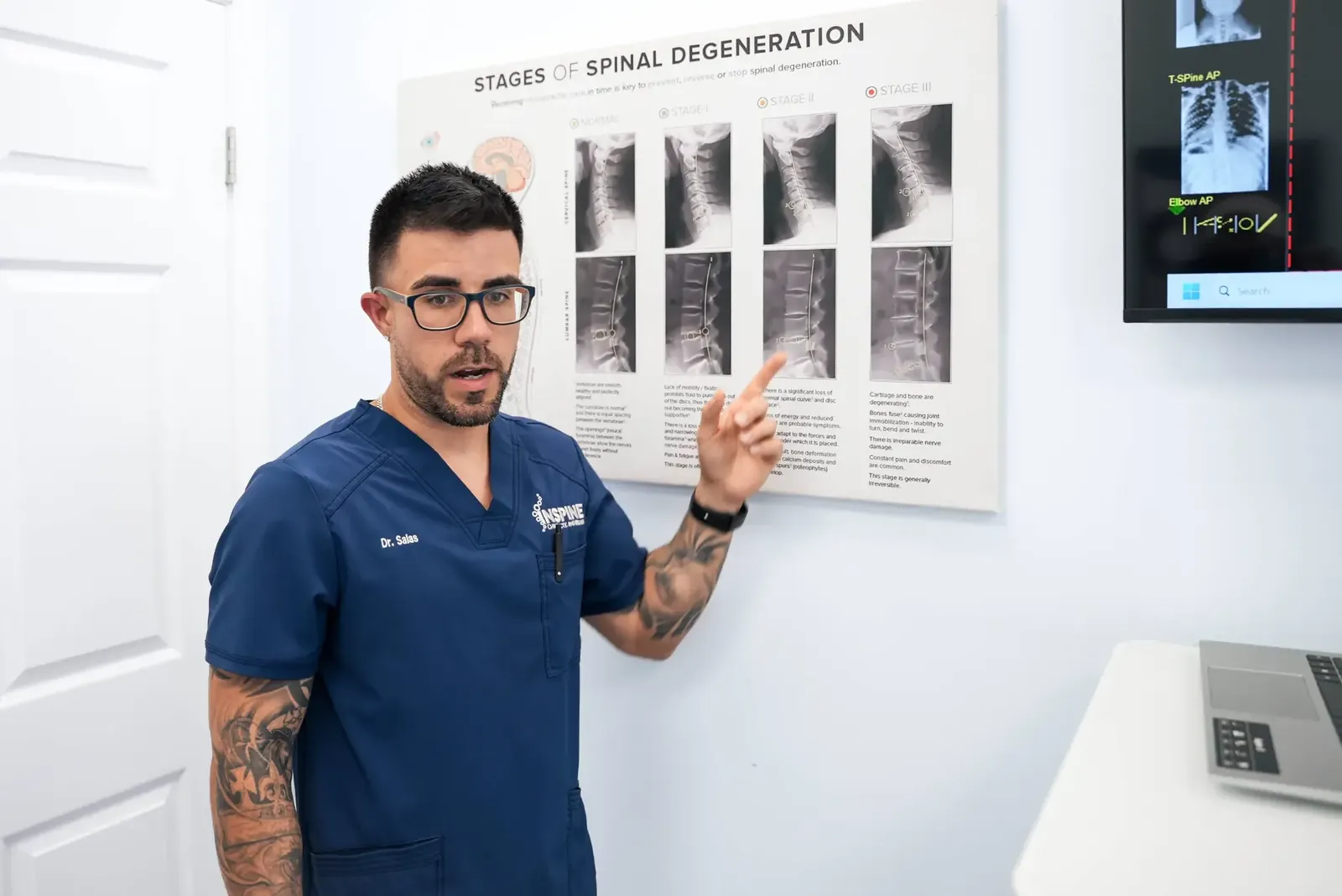 A chiropractor pointing to a spinal degeneration chart on the treatment room wall.