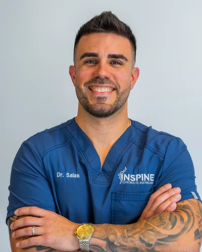Dr. Edgardo Salas, co-owner and Doctor of Chiropractic at Inspine Chiropractic and Wellness, smiling in a blue scrub top with the clinic logo.