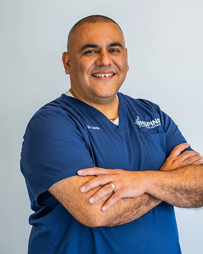 Dr. Hector Ortiz, Doctor of Chiropractic at Inspine Chiropractic, smiling in blue scrubs with crossed arms.